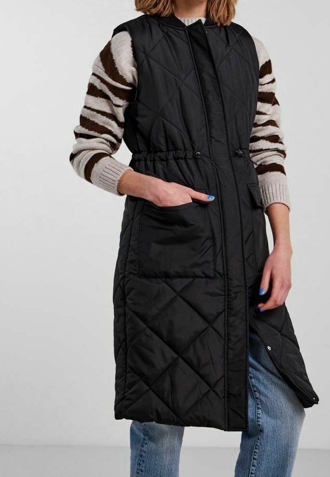 Svart vadderad ärmlös kappa med dragsko i midjan, som har två sidofickor och en fullständig dragkedja fram, bärs över en randig tröja.