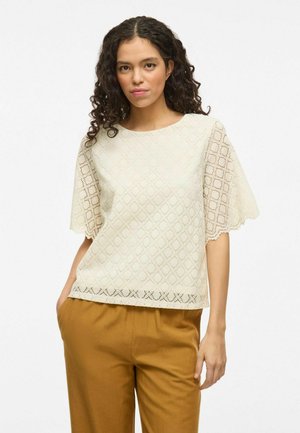 Crème lace top met diamantpatroon, geschulpte mouwen en zoom. Gecombineerd met losse bruine broeken. Textuurstof, casual ontwerp.