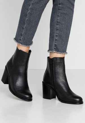 Kvinde iført sorte læder ankelstøvler med blokhæle og mørkegrå jeans med frynset kant mod enkel baggrund.