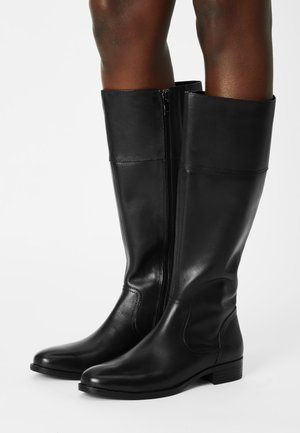 Schwarze knielange Lederstiefel mit seitlichem Reißverschluss, getragen auf nackten Beinen vor einem schlichten weißen Hintergrund.