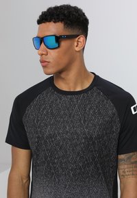Black sunglasses with blue mirrored lenses, contrasting black and gray patterned t-shirt, featuring a round neckline and short sleeves.