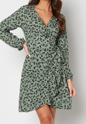 Femme en robe portefeuille verte avec motif floral noir et manches longues, debout avec une main sur la hanche.