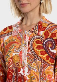 Blouse cachemire colorée en rouge, orange et bleu ; dotée d'une patte de boutonnage, d'un petit col et de motifs floraux complexes sur un tissu doux.