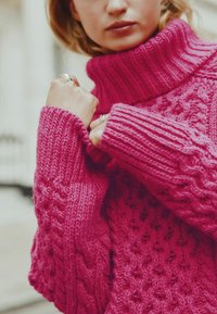 Pull en maille torsadée fuchsia avec col montant et poignets côtelés. Tissu épais et texturé avec une coupe ample, mettant en avant des motifs complexes.