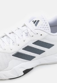 Zapatilla deportiva blanca con parte superior de malla, rayas de tela gris y cordones blancos. Cuenta con una suela texturizada con un acento negro cerca del talón.