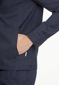 Marinblå rutmönstrad jacka i texturerat tyg med sidoficka, knappad manschett och liten varumärkesetikett på fållen.