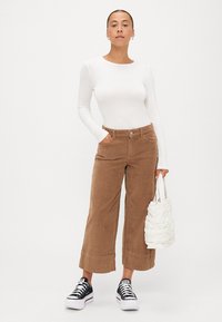 Bruine corduroy wide-leg broek, witte lange mouwen top, zwarte sneakers en een witte textuur handtas. Model staat voor een neutrale achtergrond.