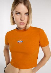 Camiseta corta naranja de algodón, con cuello redondo y un logo bordado en el centro. Manga corta y diseño ajustado.