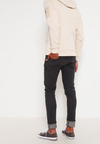 Beige hoodie met een patch op de linkermouw, gecombineerd met donkere jeans en grijze sneakers met witte zolen en opgerolde manchetten.