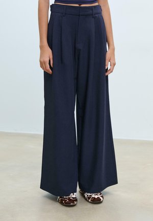 Pantaloni a gamba wide blu scuro con vita alta e dettagli plissettati, realizzati in un tessuto morbido. Abbinati a scarpe marroni con motivo.