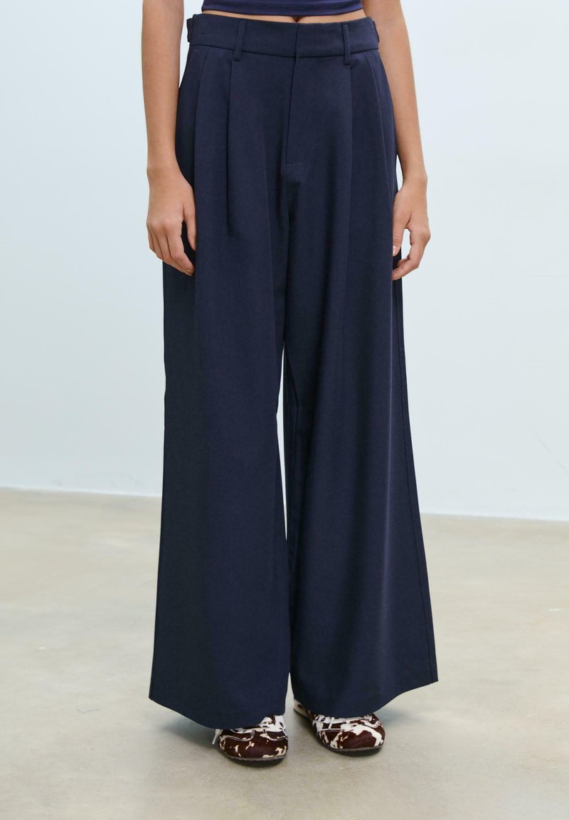 Pantaloni a gamba wide blu scuro con vita alta e dettagli plissettati, realizzati in un tessuto morbido. Abbinati a scarpe marroni con motivo.