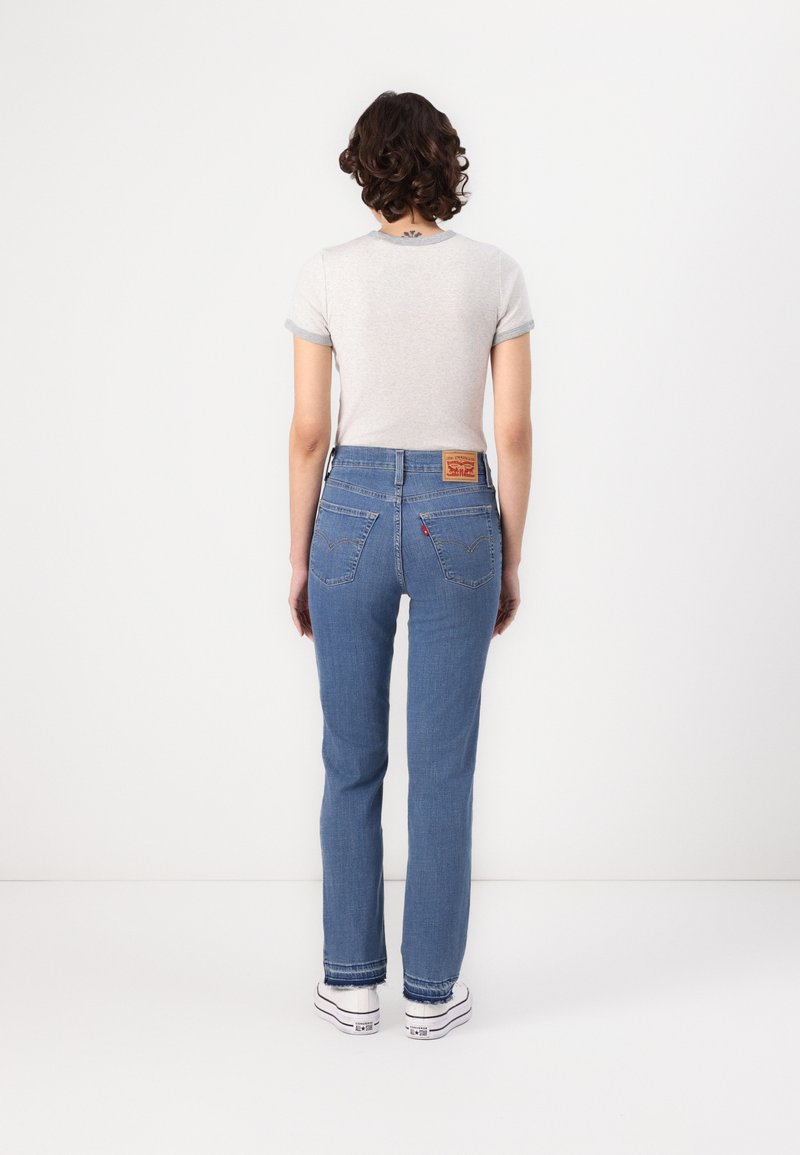 Helle graue T-Shirt mit kurzen, gerollten Ärmeln, kombiniert mit blauen, geraden Jeans. Die Jeans haben Gesäßtaschen und ein braunes Lederlogo-Label.