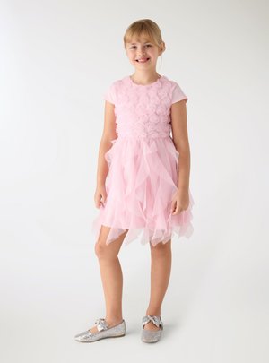 Ragazza giovane sorridente, indossa un vestito rosa con motivo floreale, gonna arricciata e scarpe glitter argento con fiocchi, in piedi su sfondo bianco.