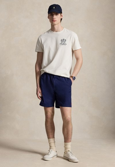 Mladý muž v námorníckej čiapke, béžovom tričku Ralph's Beach Club, námorníckych šortkách, béžových ponožkách a bielych teniskách, stojaci s jednou rukou v vrecku.