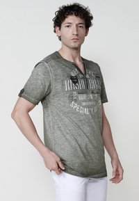 Olivinės žalios spalvos trumpomis rankovėmis t-shirt su V formos iškirpte, su spaudiniais ir siuvinėjimo detalėmis. Audinys turi dėvėtą tekstūrą.