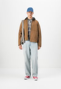 Ung mann iført blå caps, brun jakke over rutete skjorte og hettegenser, lyseblå jeans, røde joggesko, og bærer en grå skulderveske.