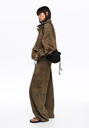 Mujer con chaqueta de ante verde oliva extragrande y pantalones anchos a juego, llevando un pequeño bolso negro de hombro y sandalias negras.