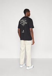 Μαύρη μπλούζα με γραφικό τύπωμα στη πλάτη, συνδυασμένη με κρεμ παντελόνι και λευκά παπούτσια. Η μπλούζα έχει κείμενο και σχέδια σε λευκό χρώμα.