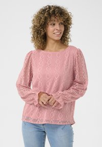 Pink blouse with textured diamond patterns, puffed sleeves, and ruffled cuffs. Worn with light blue jeans, against a white background.