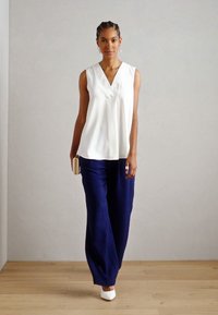Blusa bianca senza maniche con scollatura a V, abbinata a pantaloni blu navy larghi. Il modello tiene una piccola pochette e indossa tacchi bianchi.