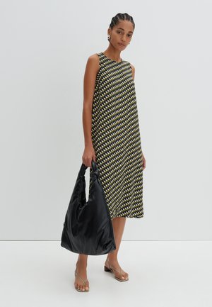 Schwarzes und gelbes gemustertes ärmelloses Kleid mit lockerer Passform und rundem Ausschnitt; hält eine große schwarze weiche Tasche; nackte Sandalen.