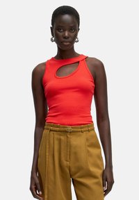 Rotes geripptes Tanktop mit einem runden Ausschnitt an der Schulter, kombiniert mit hochtaillierten, khakifarbenen, gefalteten Hosen. Glatte Stofftextur.