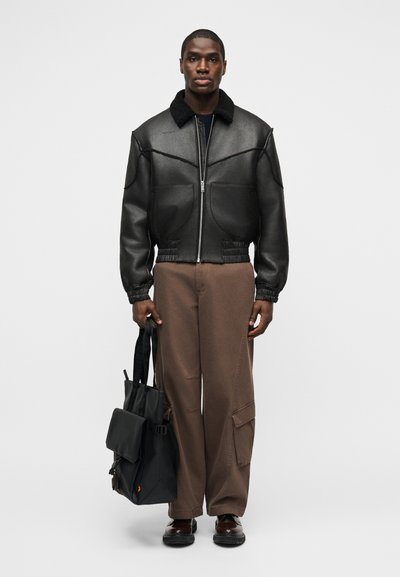 Homme portant une veste en cuir noire avec un col en peau retournée, un pantalon cargo marron, tenant un grand sac fourre-tout noir, debout devant un fond uni.