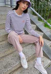 Līdzsvara krekls ar garām piedurknēm lavandas un baltā krāsā, rozā bikses un baltas kedas ar pasteļtoņiem, sēžot uz akmens pakāpieniem.