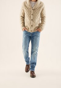 Beige Kabelstrick-Cardigan mit Schalkragen und braunen Knöpfen, kombiniert mit hellblauen Jeans und dunkelbraunen Schuhen.