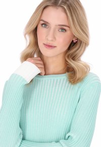 Pulover verde mentă cu dungi, cu accente albe la manșete. Modelul are păr blond ondulat și poartă cercei roz. Textură moale, design ajustat.