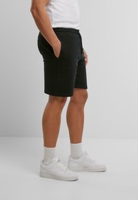 Homme portant un short noir jusqu'aux genoux, des chaussettes blanches, des baskets blanches et un t-shirt blanc, se tenant avec les mains dans les poches, vue de côté.
