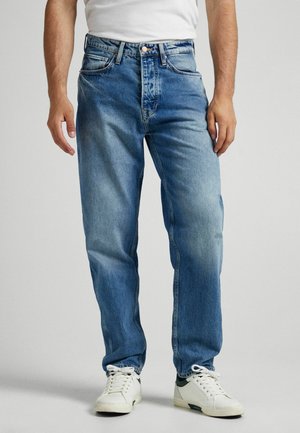 Mężczyzna w jasnoniebieskich prostych dżinsach, białych trampkach i białej koszuli, stojący na jednolitym tle.