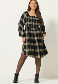 Vestido a cuadros en negro y beige con mangas largas abullonadas, escote cuadrado, corsé ajustado y falda fruncida. Llevado con botines negros.