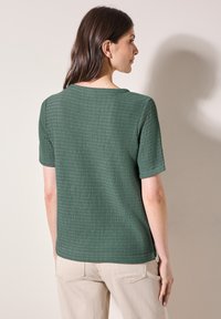 Zelený pletený top s krátkými rukávy a kulatým výstřihem, který se vyznačuje jemným rytmickým vzorem. Zadní pohled ukazuje čisté linie a švy.