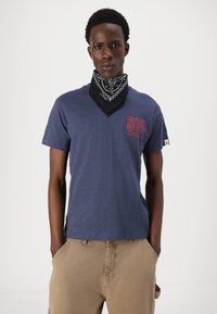 Tamsiai mėlynas medvilninis marškinėliai trumpomis rankovėmis ir V formos iškirpte, su raudona grafika; derinami su juodu bandana ant kaklo.