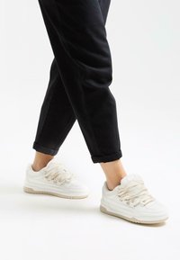 Witte sportieve schoenen met een gestructureerde mesh bovenkant, dikke zachte veters en een rubberen zool; gecombineerd met zwarte broek met opgerolde pijpen.