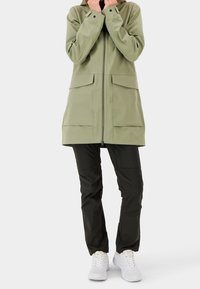 Ljust grön vattentät jacka med dragkedja fram, två stora bröstfickor och huva, kombinerad med svarta byxor och vita sneakers.