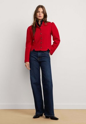 Vrouw die een rood jasje met knopen draagt over een zwart topje, met donkerblauwe wijde jeans en zwarte puntige schoenen, staand tegen een effen achtergrond.