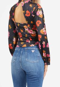 Blouse fleurie manches longues avec une découpe dans le dos, ornée de fleurs orange vif et roses sur un fond noir, associée à un jean bleu ajusté.