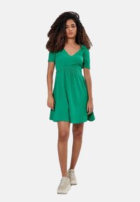 Groene jurk met korte mouwen en V-halslijn, gathered taille, vloeiende rok en een gestructureerde stof; gecombineerd met beige sneakers.