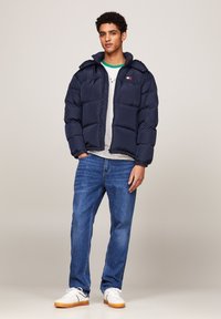 Giubbotto imbottito blu con design trapuntato, polsini elasticizzati e cappuccio. Indossato sopra una maglietta grigia chiara, abbinato a jeans blu e sneakers bianche.