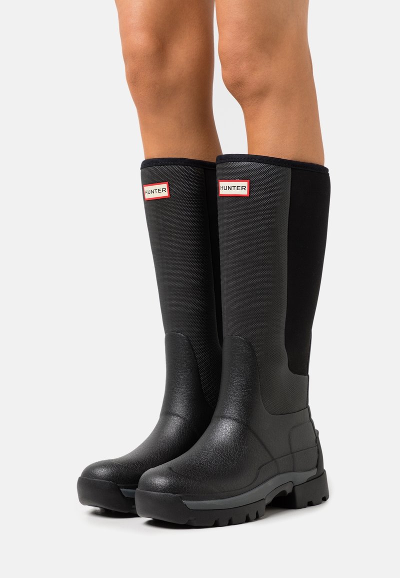 Bottes noires en caoutchouc, montantes jusqu'au genou, avec un design de panneau texturé. Elles présentent des semelles prononcées et un bout arrondi. Le logo de la marque est affiché de manière proéminente.