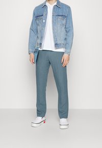 Hellblaue Jeansjacke, weißes T-Shirt und hellblaue Hose. Person trägt weiße Slipper mit roten Akzenten und hält eine blaue Tasche.