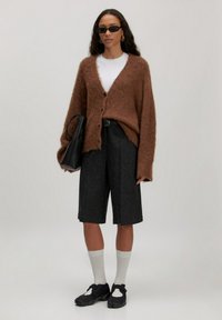 Bruine fluffy cardigan, wit t-shirt, zwarte gestructureerde shorts, witte sokken, zwarte schoenen met bandjes, dragend een zwarte tas; schone achtergrond.