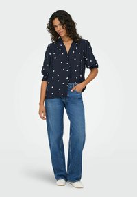 Marineblå skjorte med knapper og korte, puffede ærmer, prydet med hvide hjerte-mønstre; sat sammen med lyseblå vide jeans og hvide sneakers.