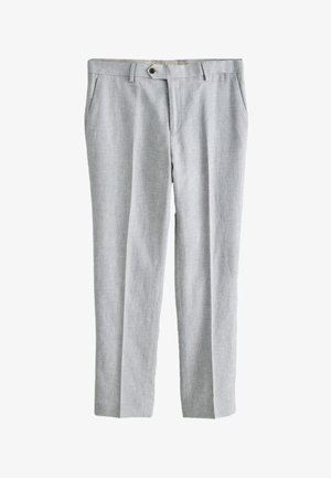 Mężczyzna stoi z jedną ręką w kieszeni, ubrany w jasnoszary garnitur, białą koszulę i czarne eleganckie buty na jednolitym beżowym tle.