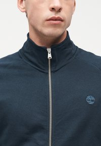 Άνδρας που φοράει ναυτικό μπουφάν με φερμουάρ, ρηχτό γιακά και μικρό κεντημένο λογότυπο δέντρου στο αριστερό στήθος.