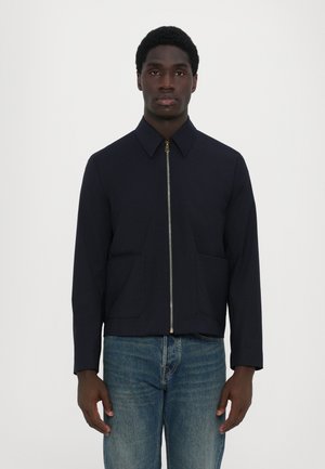 Marinefarvet jakke med lynlås foran, spidse kraver og to forlommer. Har en tætsiddende design og glat tekstur. Bæres over blå jeans.