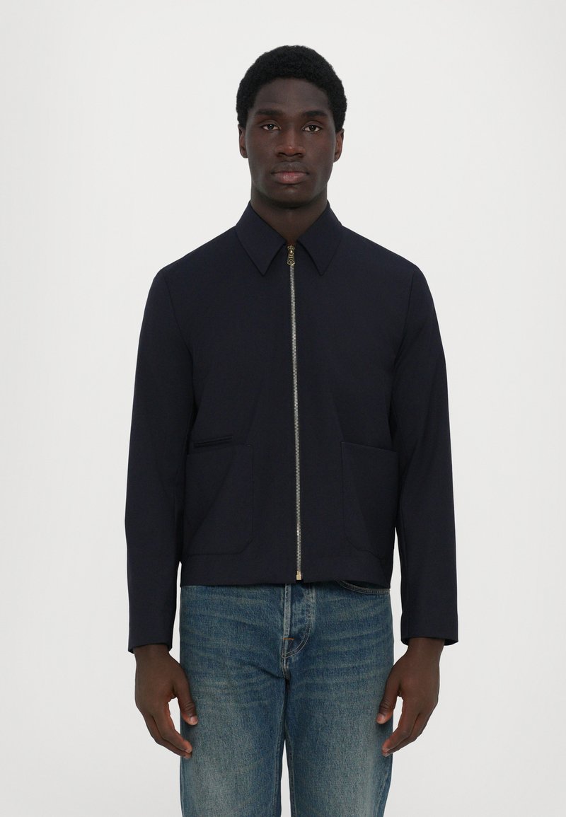 Giacca navy con zip frontale, colletto a punta e due tasche frontali. Caratterizzata da un design aderente e da una texture liscia. Indossata sopra un paio di jeans blu.