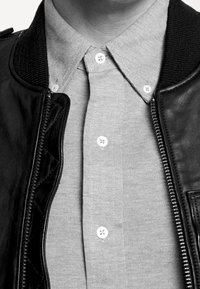 Svart läderjacka med ribbad krage och dragkedja. Grå skjorta med krage under, med vita knappar och en texturerad tyg.